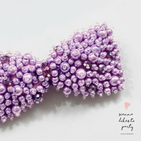 Close up of designer childrens lavender purple beaded hair bow hair clip embellished with pearl beads and small rhinestones, shown on a clean white background with a 'sienna likes to party' watermark in the lower right.