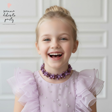 Young girl wearing a purple dress with ruffled sleeves and a matching handmade couture lilac and amethyst choker necklace by Sienna Likes to Party, smiling against a white background.